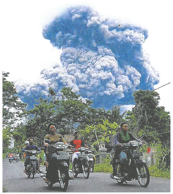 巨大的火山灰雲在樹木後方升起,前方道路上有人騎摩托車的照片